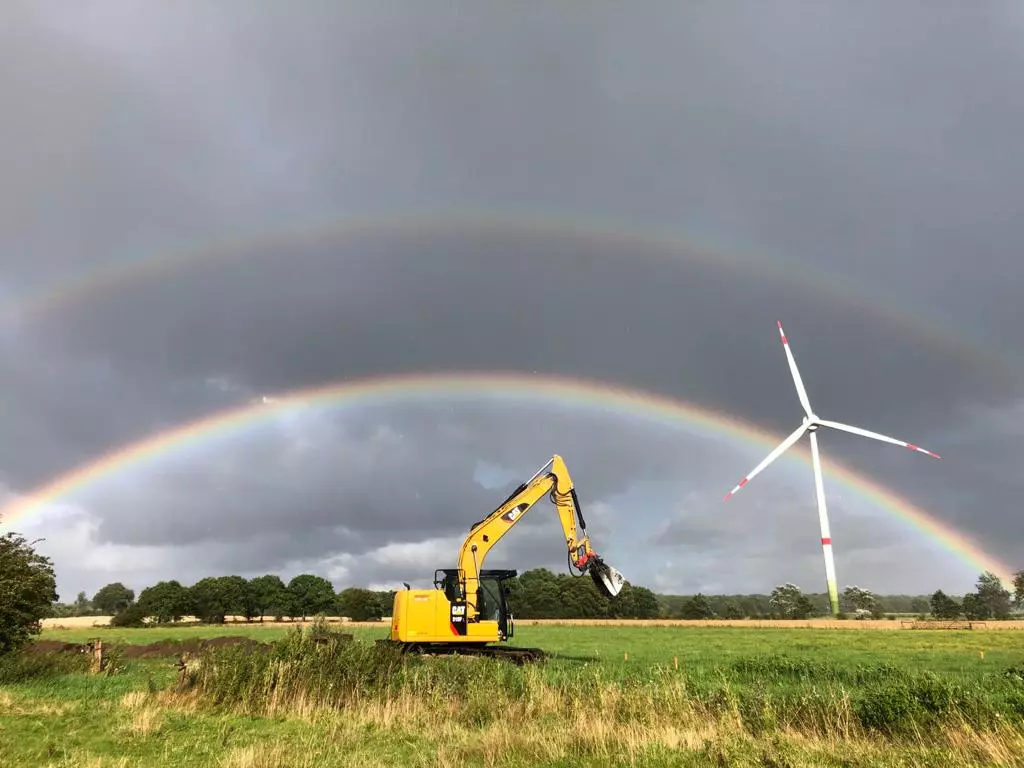 Bagger der Uwe Schröder Erdbau GmbH auf einem Feld mit Regenbogen und Windkraftanlage im Hintergrund