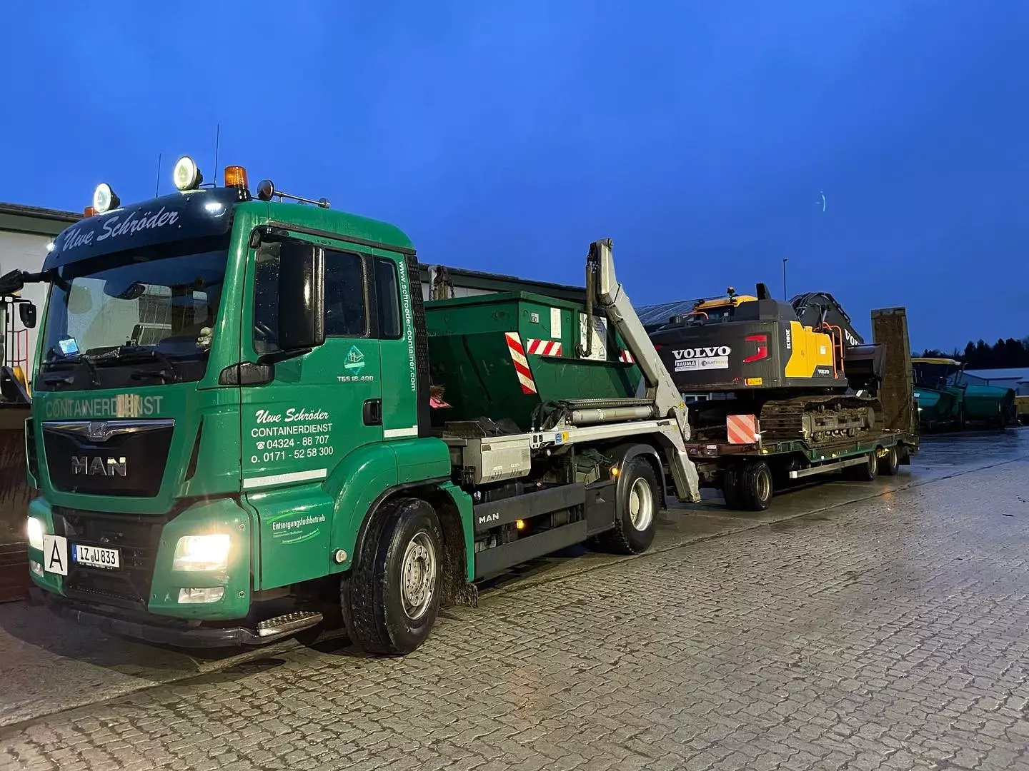 MAN Containerdienst-LKW und Tieflader mit Bagger der Uwe Schröder Erdbau GmbH bei Transporten in der Dämmerung